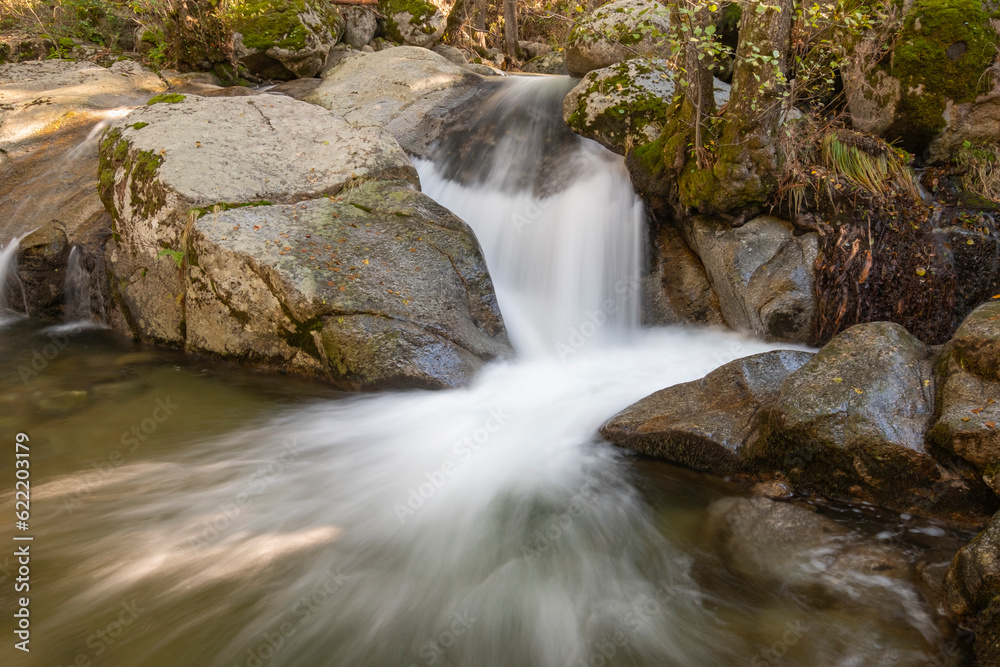 Fototapeta premium Small waterfall in a river whose flow runs between large granite stones deep in a dark forest. Concept of peace and relaxation, water stream and silk water.