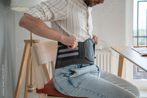 Man sitting on chair wearing back brace to reduce hernias, using decompression corset for herniated discs at home, relieving symptoms of degenerative disc disorder. Recovery after lower back surgery