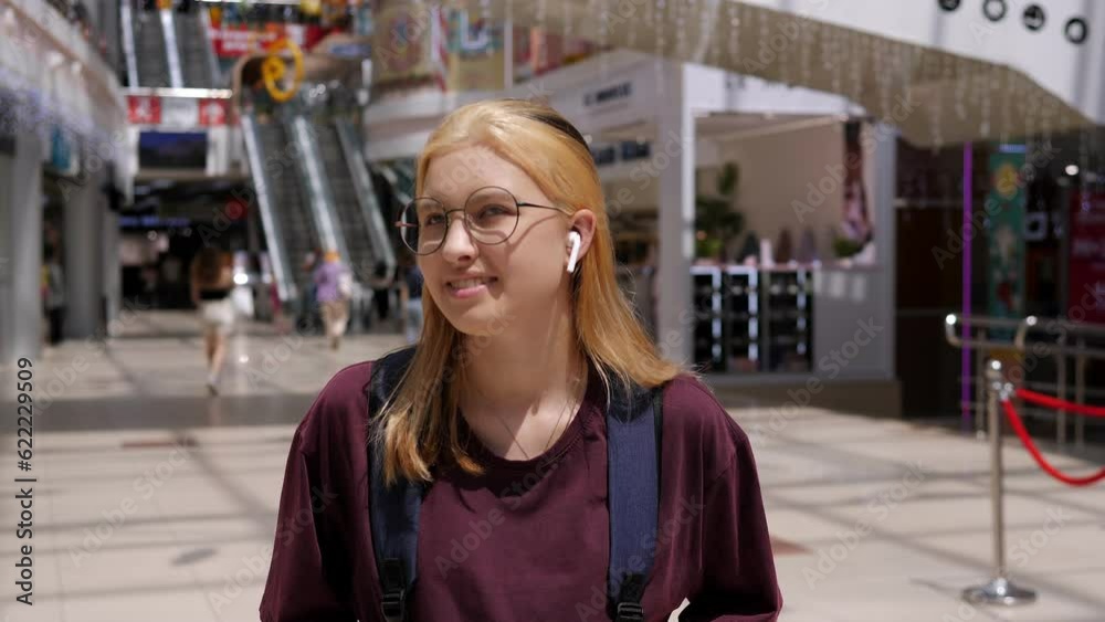 A stylish teenage girl in round glasses and wireless headphones with a backpack is standing in a shopping mall, with an escalator in the background. A stylish teenage girl in round glasses and wireless headphones with a backpack is standing in a shopping mall, with an escalator in the background.