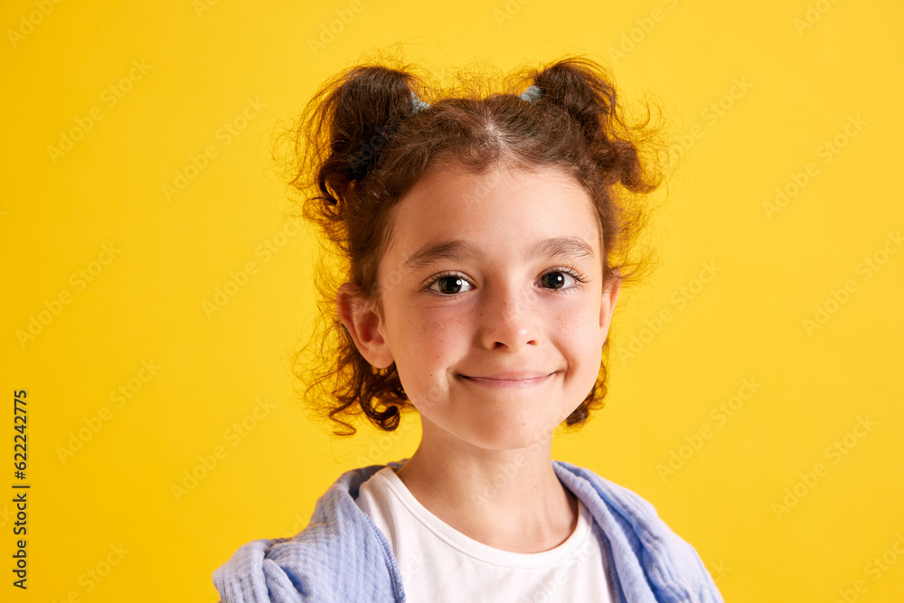 Close-up portrait of beautiful little girl, child with curly hair posing, looking at camera, smiling against yellow studio background. Concept of emotions, childhood, education, fashion, lifestyle, ad Close-up portrait of beautiful little girl, child with curly hair posing, looking at camera, smiling against yellow studio background. Concept of emotions, childhood, education, fashion, lifestyle, ad