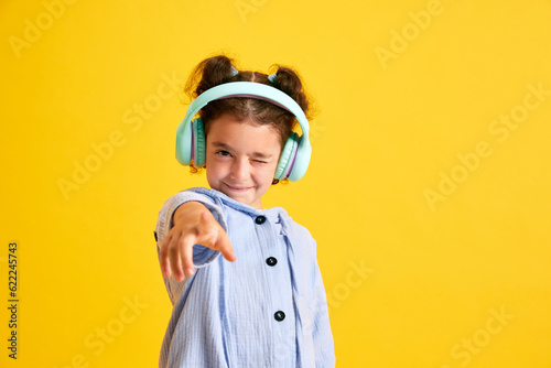 Portrait of positive little girl child listening to music in headphones and grimacing against yellow studio background. Concept of emotions  childhood  education  fashion  lifestyle  ad