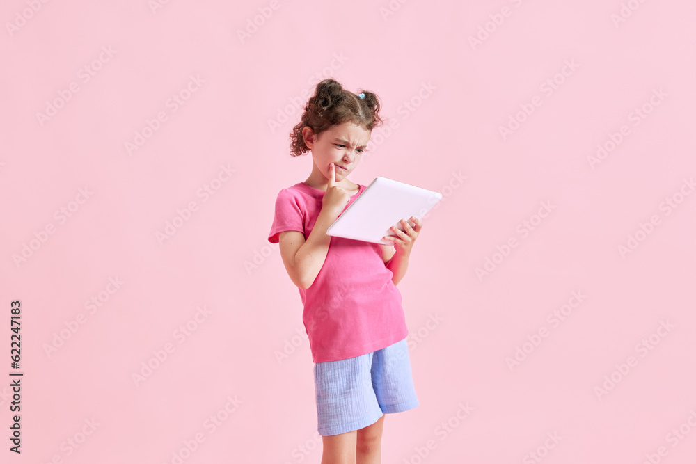 Portrait of child, little girl looking in tablet with thoughtful, concentrated face against pink studio background. Online lesson. Concept of emotions, childhood, education, fashion, lifestyle, ad Portrait of child, little girl looking in tablet with thoughtful, concentrated face against pink studio background. Online lesson. Concept of emotions, childhood, education, fashion, lifestyle, ad