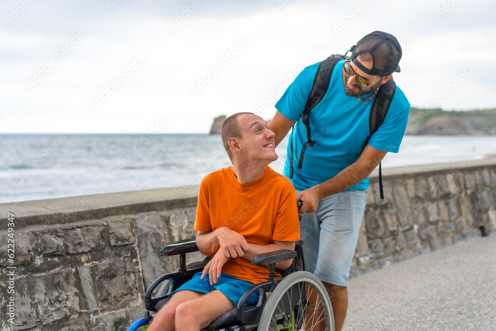 Disabled person in a wheelchair being pushed by a friend on the beach promenade, having fun in summer Disabled person in a wheelchair being pushed by a friend on the beach promenade, having fun in summer