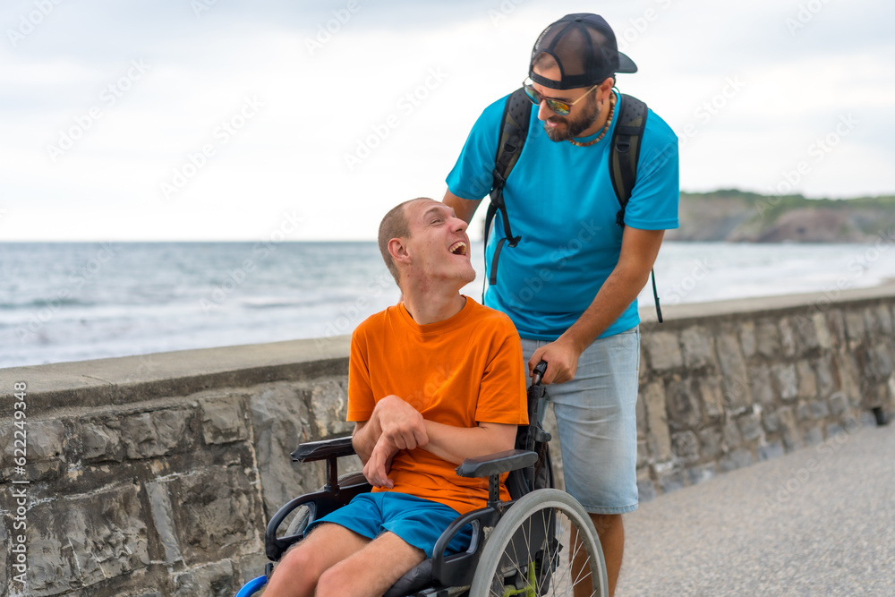 Disabled person in a wheelchair being pushed by a friend on the beach promenade, having fun in summer Disabled person in a wheelchair being pushed by a friend on the beach promenade, having fun in summer