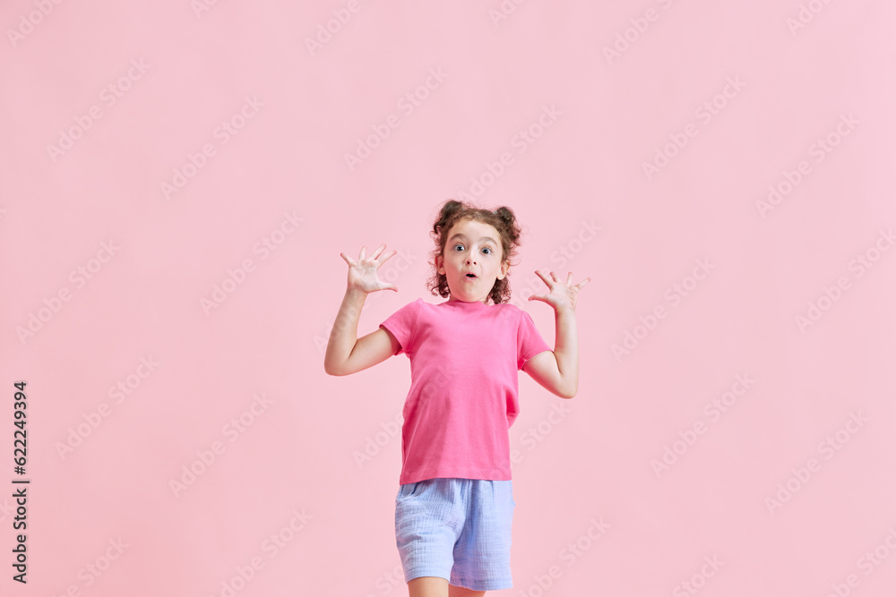 Portrait of little emotional girl, child expressing excitement and surprise against pink studio background. Concept of emotions, childhood, education, fashion, lifestyle, ad