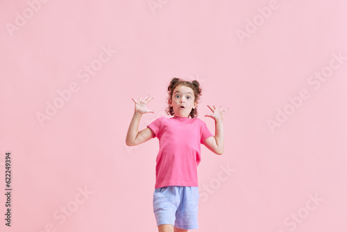 Portrait of little emotional girl, child expressing excitement and surprise against pink studio background. Concept of emotions, childhood, education, fashion, lifestyle, ad