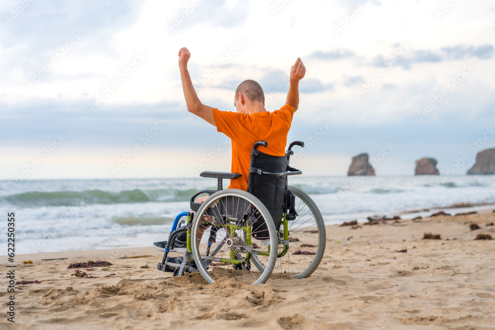 Fototapeta premium A disabled person on his back in a wheelchair on the beach with open arms enjoying the freedom of the sea and nature