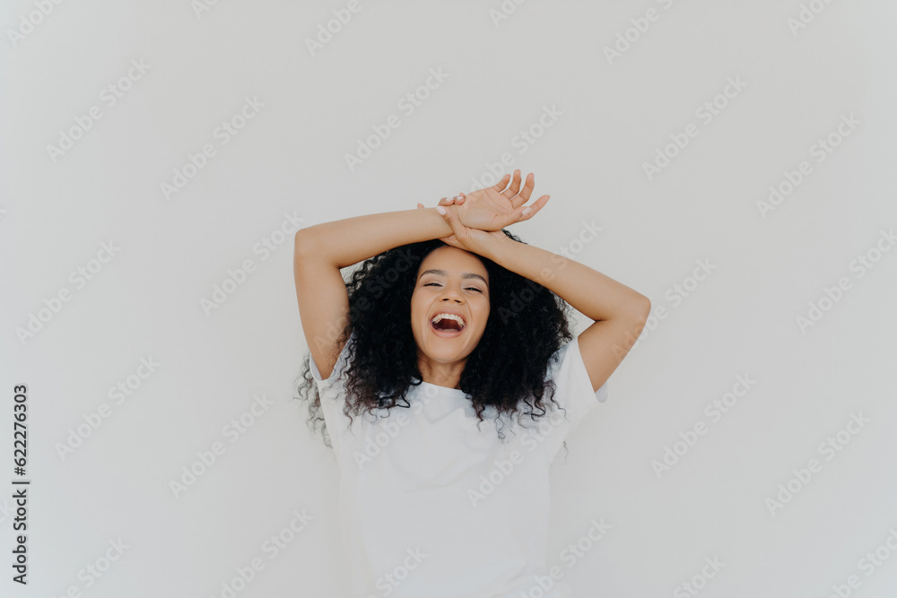 Fototapeta premium Overjoyed curly-haired lady, hands on forehead, laughing with mouth wide open, energetic vibes. Casual attire, white studio backdrop. Hilarious joke!