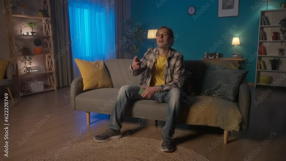 A man sits on the couch turns on the TV with the remote control. A man adjusts the brightness of the lighting in a room using a smart home control app on a smartphone. HDR BT2020 HLG Material. A man sits on the couch turns on the TV with the remote control. A man adjusts the brightness of the lighting in a room using a smart home control app on a smartphone. HDR BT2020 HLG Material.
