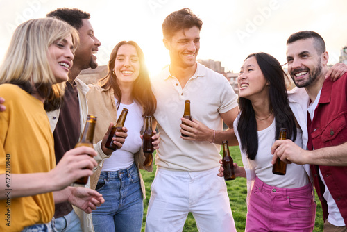 Multi-ethnic friends chatting over a bottle of beer outdoors. Group of young diverse people standing party drinking bottled beer talking and laughing in city park. Concept of hanging out in community 