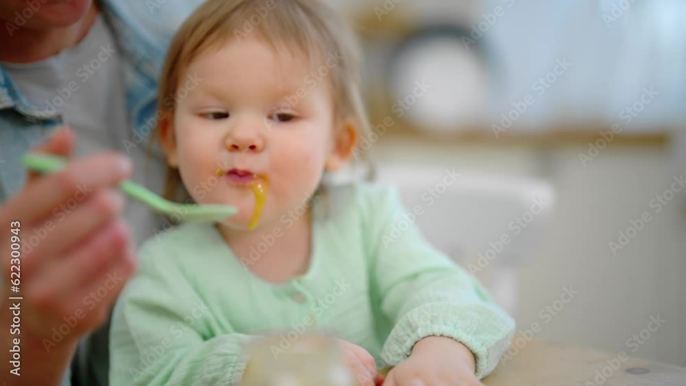 Mom feeds her baby playing with food. Toddler eats fruit or vegetable puree. Mother holds child on knees at table on kitchen. Raising child, kid's nutrition, motherhood, love care in family concept. Mom feeds her baby playing with food. Toddler eats fruit or vegetable puree. Mother holds child on knees at table on kitchen. Raising child, kid's nutrition, motherhood, love care in family concept.