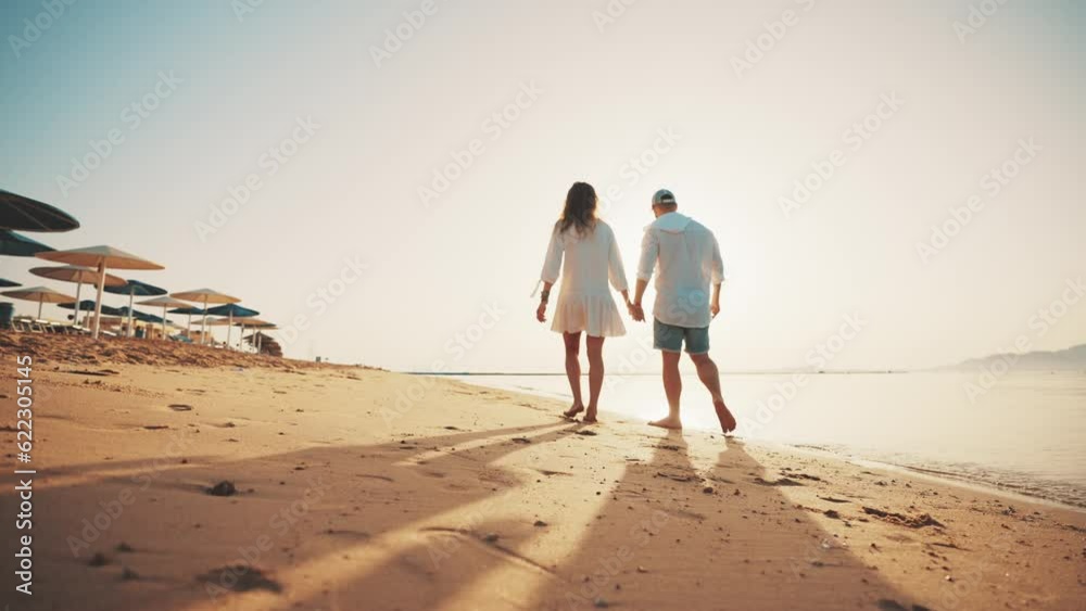 Loving couple walking along sandy ocean beach at sunny day talking enjoying time together. Man woman having romantic date, honeymoon near sea, back view. Tourism, travel family holidays concept.