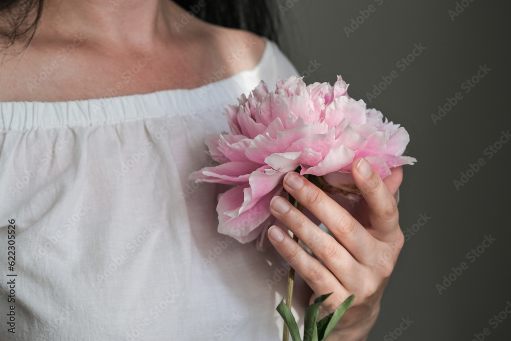 A young woman in a white dress tenderly holds a pink peony in her hands.