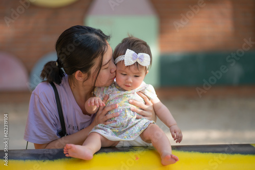 outdoors lifestyle portrait of Asian Chinese woman playing with beautiful and adorable baby girl smiling cheerful on playground playing on swing at city park wearing a cute ribbon