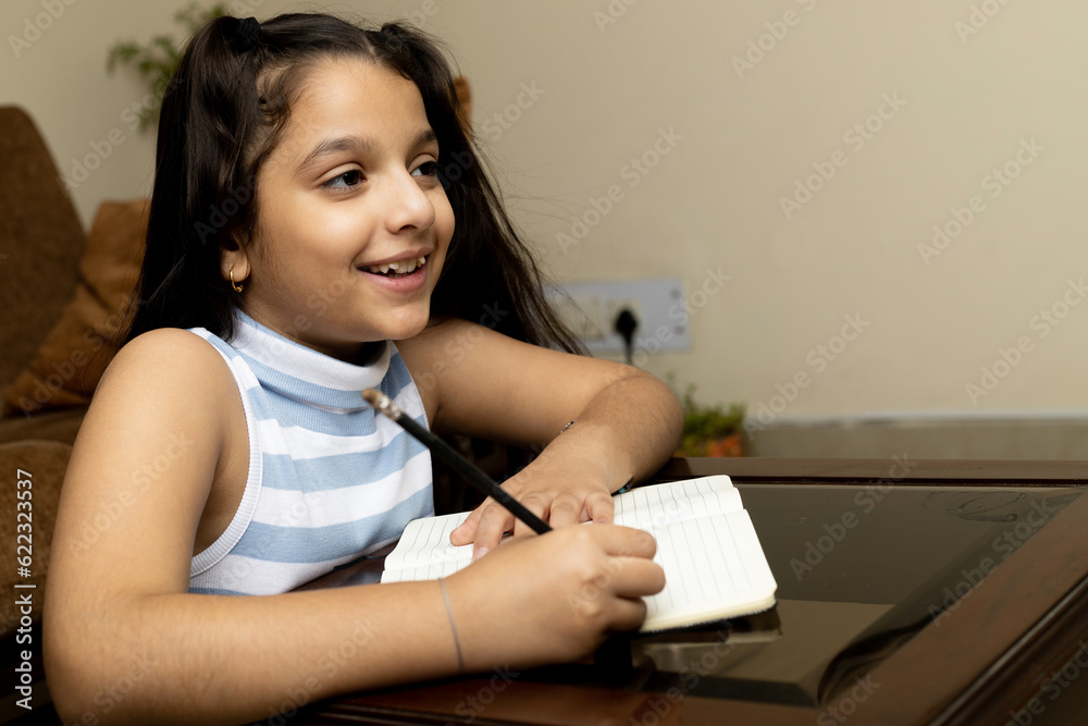 Happy cute Indian preteen kid girl student studying at table in home, looking in Tablet doing online studies Happy cute Indian preteen kid girl student studying at table in home, looking in Tablet doing online studies