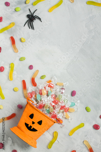Halloween orange bucket with face, with candies inside, jujube and rubber spider on gray concrete background