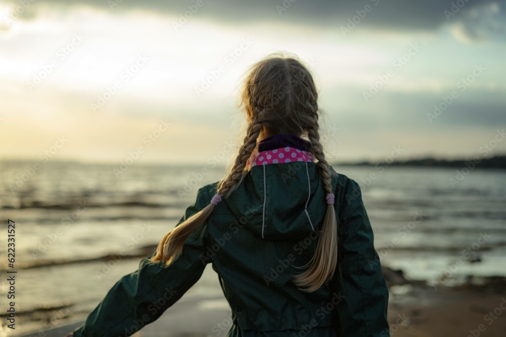 back view of caucasian blond girl with hairs styled with two plaits looking at sea on sunset