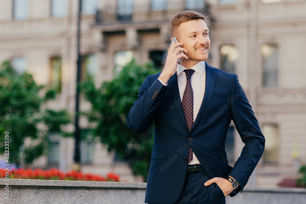 Positive successful businessman shares news with friends, glad to accomplish new innovative project, stands outdoor, keeps hand in pocket, dressed formally, notices something into distance aside Positive successful businessman shares news with friends, glad to accomplish new innovative project, stands outdoor, keeps hand in pocket, dressed formally, notices something into distance aside