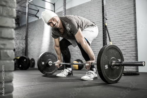 Smiling guy with a beard prepares to raise up a barbell in the gym on the gray brick wall background. He wears sportswear, white sneakers and a white cap. Horizontal.