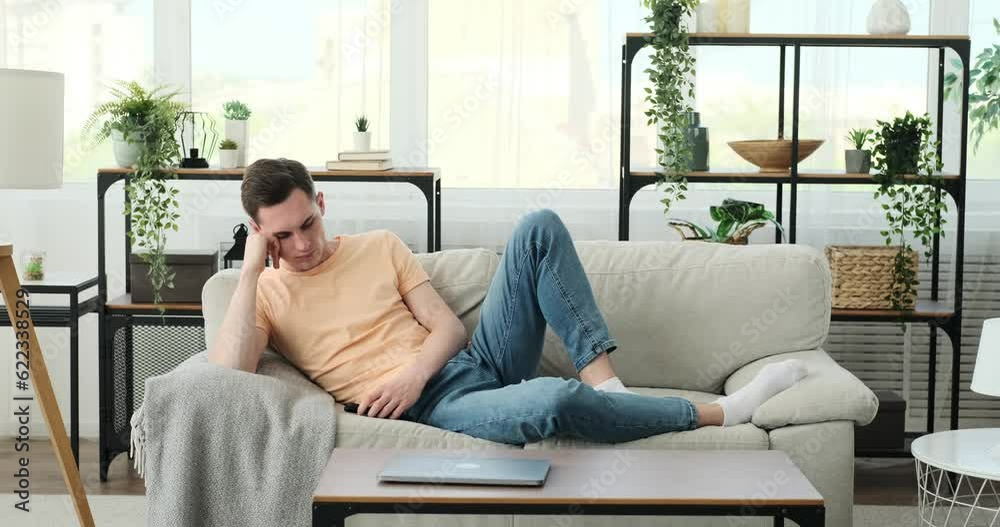 Caucasian man as he finds himself trapped in the doldrums of monotony at home. Seated on the sofa, his tired gaze and slouched posture reflect his longing for excitement and stimulation. Caucasian man as he finds himself trapped in the doldrums of monotony at home. Seated on the sofa, his tired gaze and slouched posture reflect his longing for excitement and stimulation.