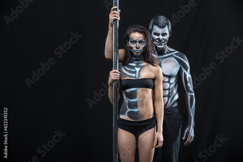 Grinning couple of pole dancers with horrific body-art stands next to a pylon on a dark background in the studio. They dressed in black sportswear and hold the right hands on the pylon. Horizontal. © Designpics