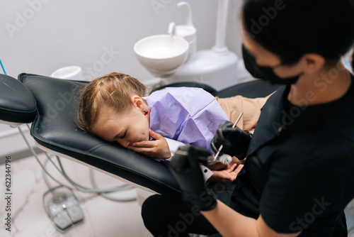 Top view of frightened little child girl sitting in dental chair being scared and turning head away from medical equipment held by experienced female dentist. Concept of children teeth treatment