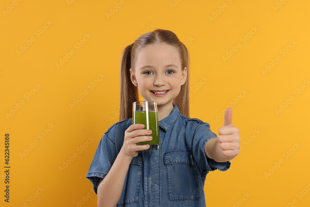Cute little girl with glass of fresh juice showing thumbs up on orange background Cute little girl with glass of fresh juice showing thumbs up on orange background