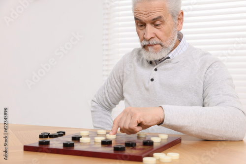 Playing checkers. Concentrated senior man thinking about next move at table in room