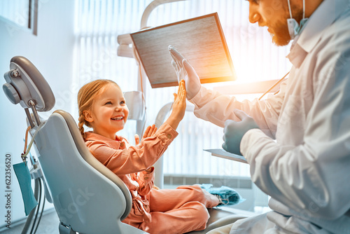 A little girl is sitting in a dentist's chair, giving a high five to the doctor and laughing. Dental care, trust and patient care. Children's dentistry.Sunlight.