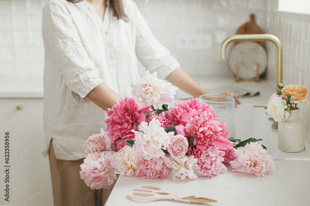 Woman arranging beautiful peonies in vase at sink with brass faucet in new modern kitchen. Female decorating flowers on background of granite countertop, hands with flowers close up Woman arranging beautiful peonies in vase at sink with brass faucet in new modern kitchen. Female decorating flowers on background of granite countertop, hands with flowers close up