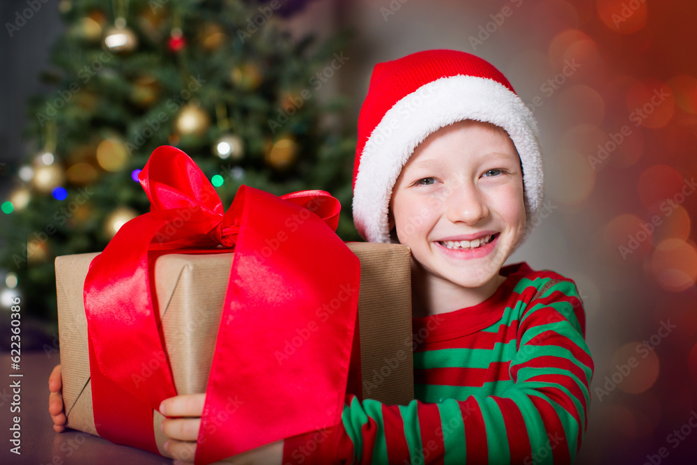 smiling happy boy in elf hat holding huge christmas present by the tree waiting for a miracle at holiday time smiling happy boy in elf hat holding huge christmas present by the tree waiting for a miracle at holiday time