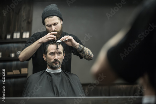 Careful barber with a big beard and a tattoo is combing the hair of the bearded man in the black hair cutting cape in the barbershop. He also fixes client's strands with the hairgrips. Horizontal.