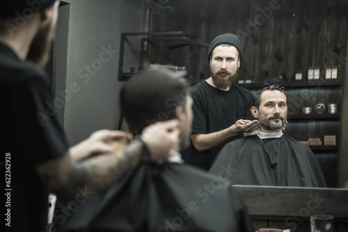 Man and a barber reflected in a mirror in the barbershop. Hairdresser wears a black T-shirt with a cap and has a hair comb. Client wears a black cutting hair cape and has hairgrips on his head. photo