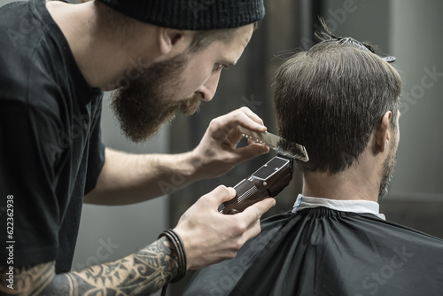 Incomparable barber with a beard and a tattoo is cutting the hair of his client in the black cape in the barbershop. He is using a cutting comb and a hair clipper. Hairdresser dressed in black wear.