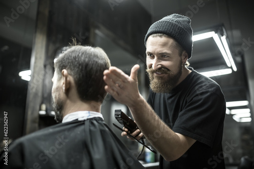 Happy barber with a beard looks at the hair of his client in the barbershop. He holds a cutting comb and a hair clipper. Hairdresser wears a black T-shirt and a cap. Horizontal.
