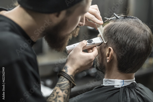 Nonpareil barber with a beard and a tattoo is cutting the hair of his bearded client in the barbershop. He is using a cutting comb and a hair clipper. Customer has hairgrips on the head. Horizontal.