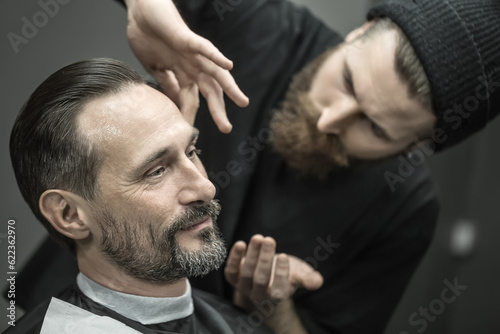 Joyful man with a beard in the barbershop. Bearded barber in a black T-shirt and a cap applies a shaving gel on client's skin. Closeup. Horizontal.