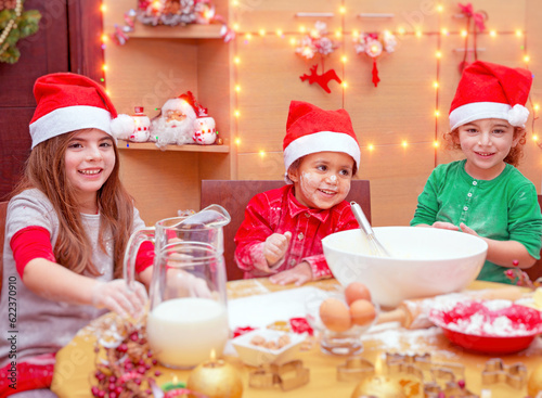 Happy children making Christmas cookies at home, three cute little kids wearing red Santa hats preparing festive tasty sweets, traditional celebration of winter holidays