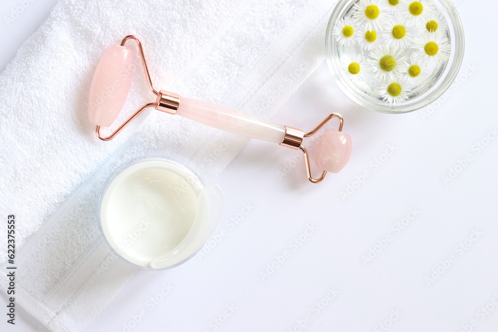 Fototapeta premium Rose quartz facial roller. Facial massage roller, chamomile water, cream and white towel. Flat lay on a light background. Top view, copy space. Spa