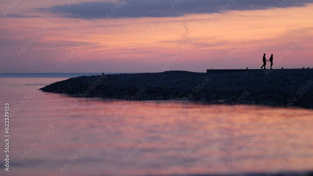 A couple of lovers standing kiss on the pier against the background of the evening sky. Silhouettes of unrecognizable people. Sunset on the sea. Romantic evening. Light waves splash near the shore. A couple of lovers standing kiss on the pier against the background of the evening sky. Silhouettes of unrecognizable people. Sunset on the sea. Romantic evening. Light waves splash near the shore.