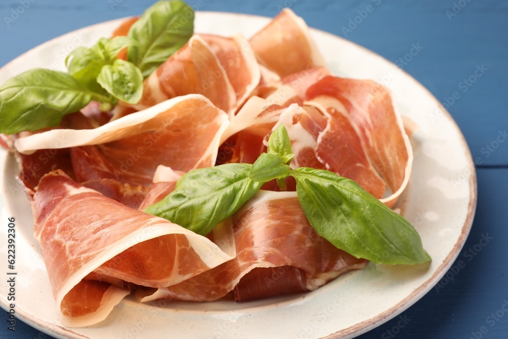 Slices of tasty cured ham and basil on blue wooden table, closeup
