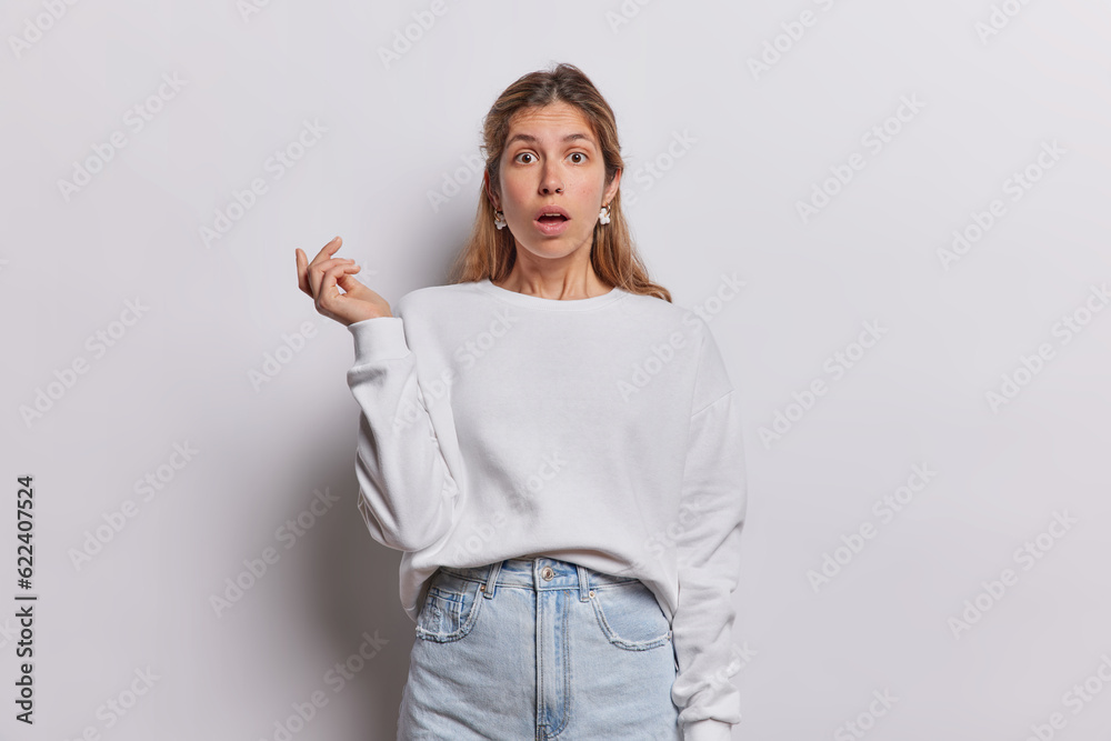Photo of surprised European woman gasps from wonder and big shock hears unbelievable news holds breath dressed in casual pullover and jeans isolated over white background. Human reactions concept
