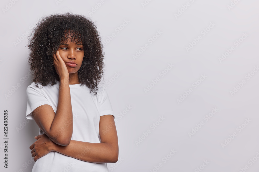 Photo of dissatisfied curly haired woman keeps hand on cheek concentrated aside dressed in casual t shirt isolated over white background copy space for your promotional content or advertisement Photo of dissatisfied curly haired woman keeps hand on cheek concentrated aside dressed in casual t shirt isolated over white background copy space for your promotional content or advertisement