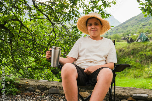 A boy in a straw hat relaxes on a tourist chair under a tree and drink tea from thermocup on background of mountains. Concept of tourism. photo