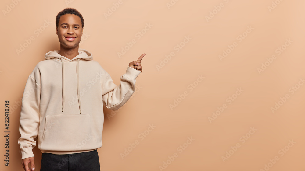 Studio shot of handsome dark skinned man with short hair points index finger aside on blank space advertises product dressed in hoodie isolated over brown background. Check this awesome promo Studio shot of handsome dark skinned man with short hair points index finger aside on blank space advertises product dressed in hoodie isolated over brown background. Check this awesome promo