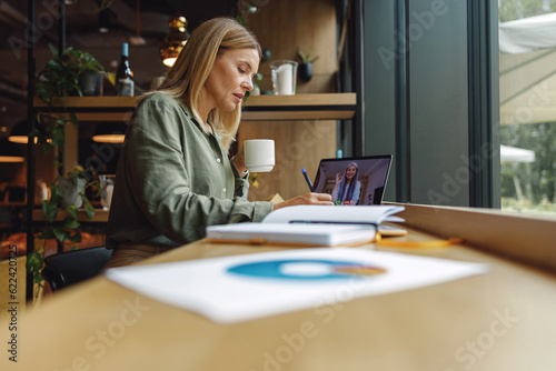 Middle-aged businesswoman working on laptop and making notes in cozy cafe while drinking coffee