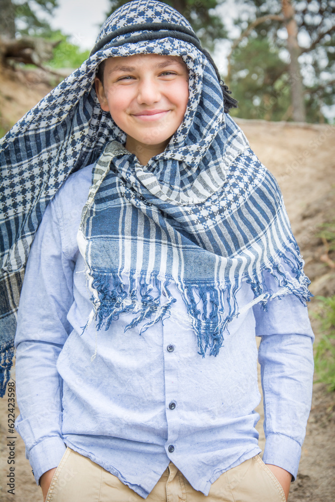 In the summer, a beautiful boy in a arafatka, an Arab scarf, stands on the road in the forest. In the summer, a beautiful boy in a arafatka, an Arab scarf, stands on the road in the forest.