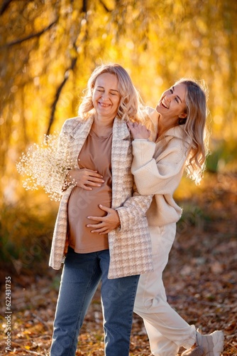 Adult daughter hugs her pregnant mom while standing in autumn park