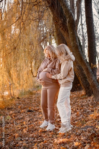 Adult daughter hugs her pregnant mom while standing in autumn park photo