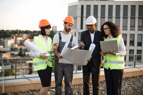 Group of four multiracial people in suits and helmets brainstorming for common project of new real estate building. Construction team working with blueprints and modern laptop in fresh air.